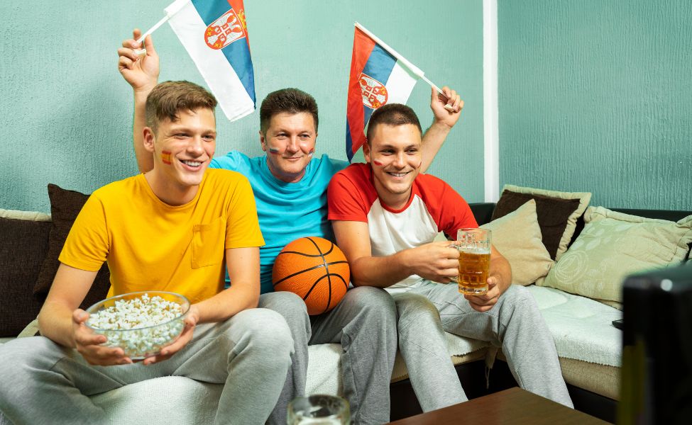 Three people holding popcorn, cheering and holding Serbian flags 